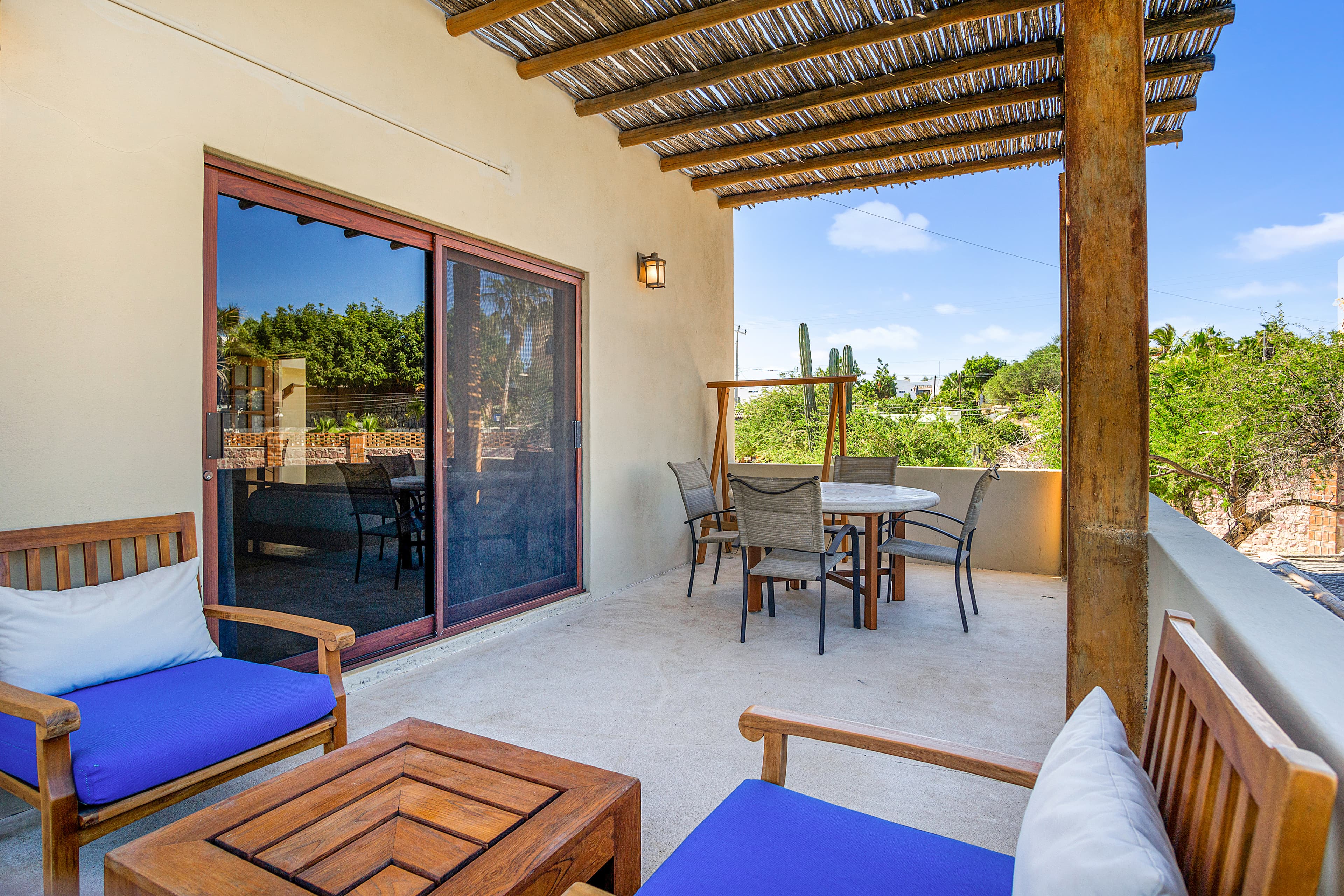 A cozy outdoor terrace featuring wooden furniture and a table, surrounded by greenery under a thatched roof.