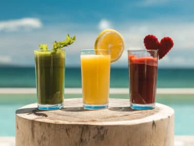 Three colorful cocktails on a wooden table with a seaside background.