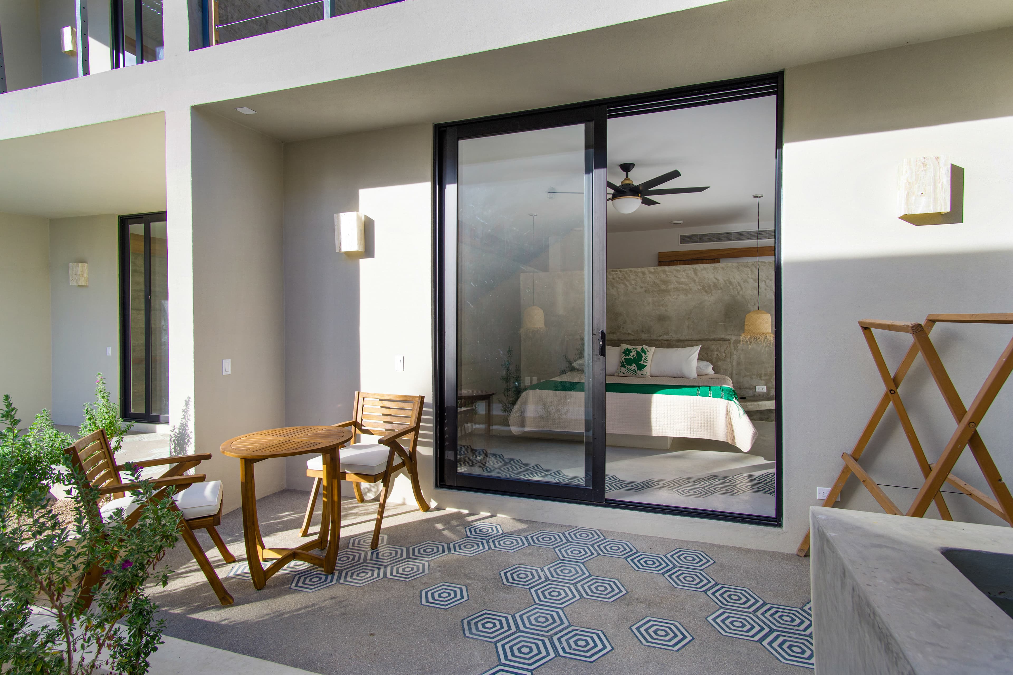 A cozy terrace with wooden furniture leads to a modern bedroom through large glass doors.
