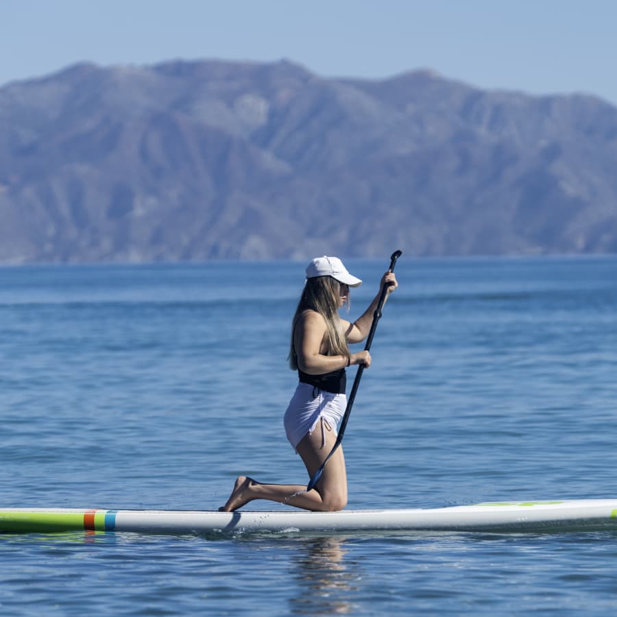 A person kneels on a paddleboard in calm water, surrounded by mountains.