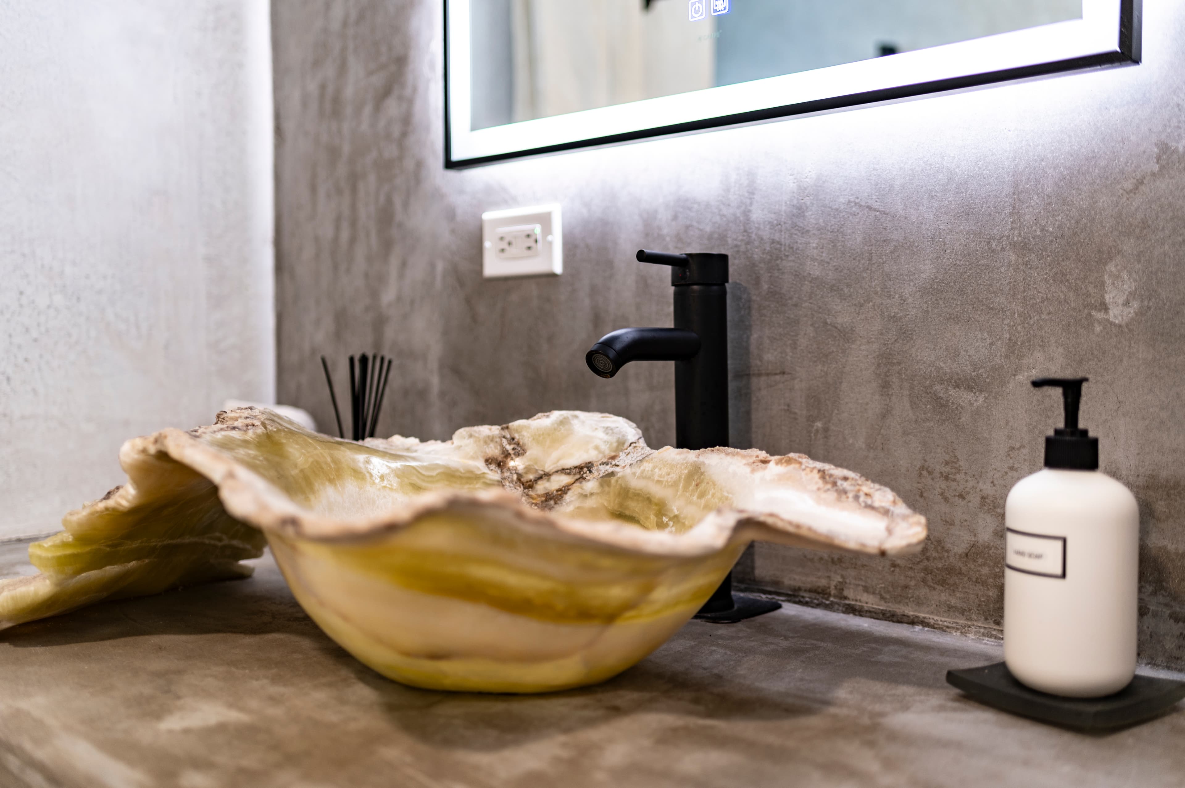 A unique, shell-shaped sink with a black faucet and a white soap dispenser on a concrete countertop.