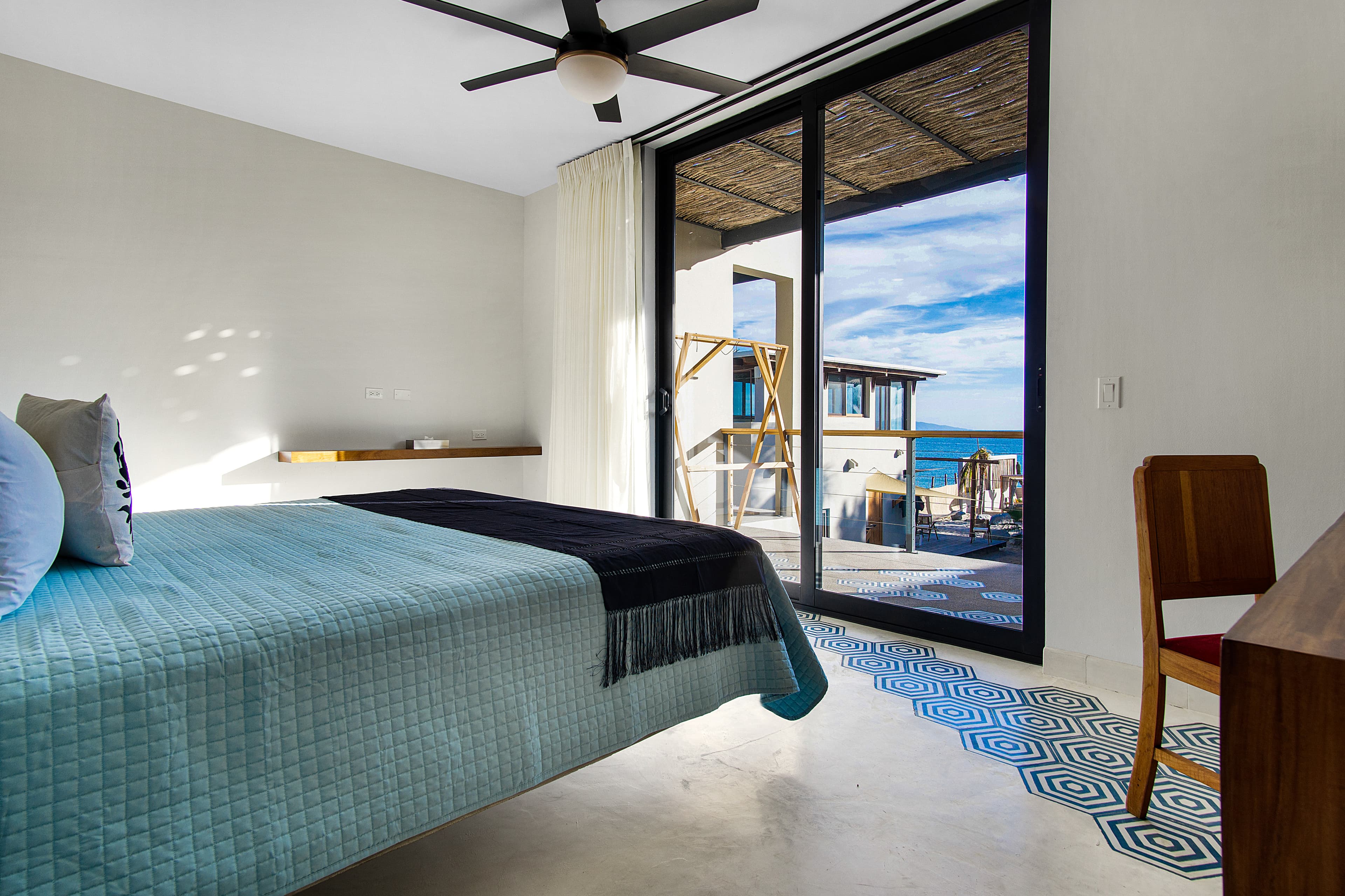 A modern bedroom with a blue quilted bedspread and large glass doors leading to a sunny terrace overlooking the ocean.