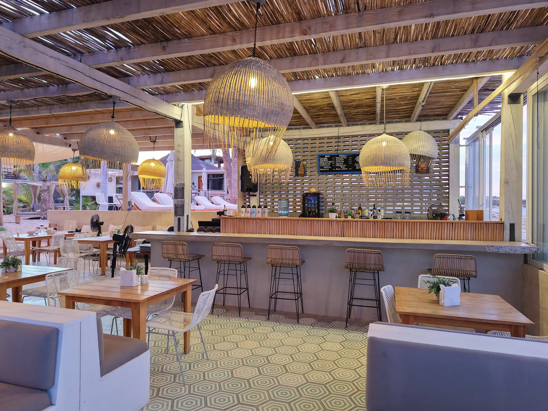 A modern restaurant interior featuring wooden tables, wicker chairs, and illuminated pendant lamps.
