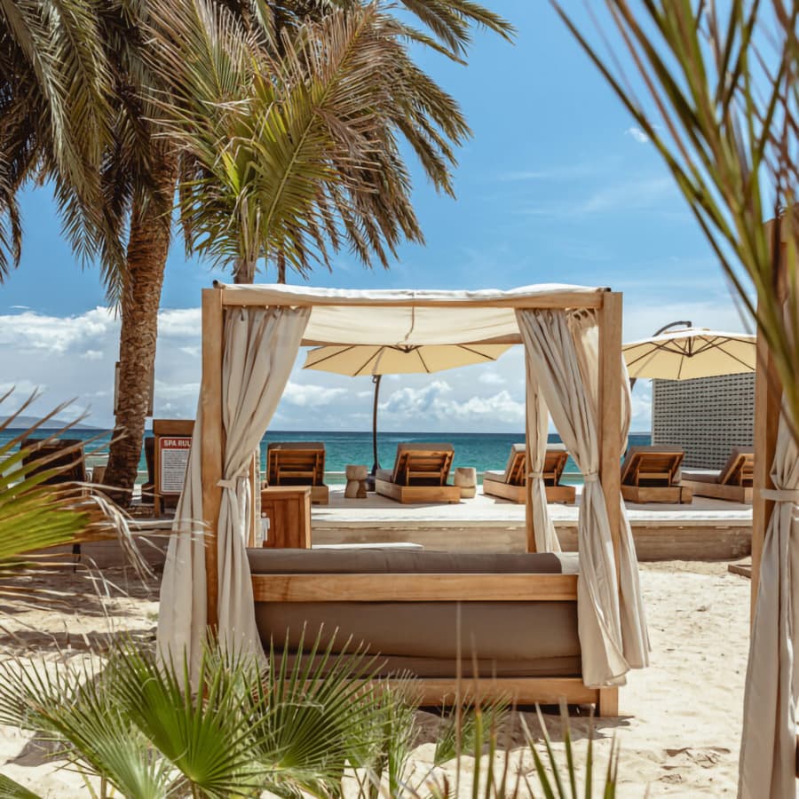 A beachside cabana surrounded by palm trees and loungers under a clear blue sky.