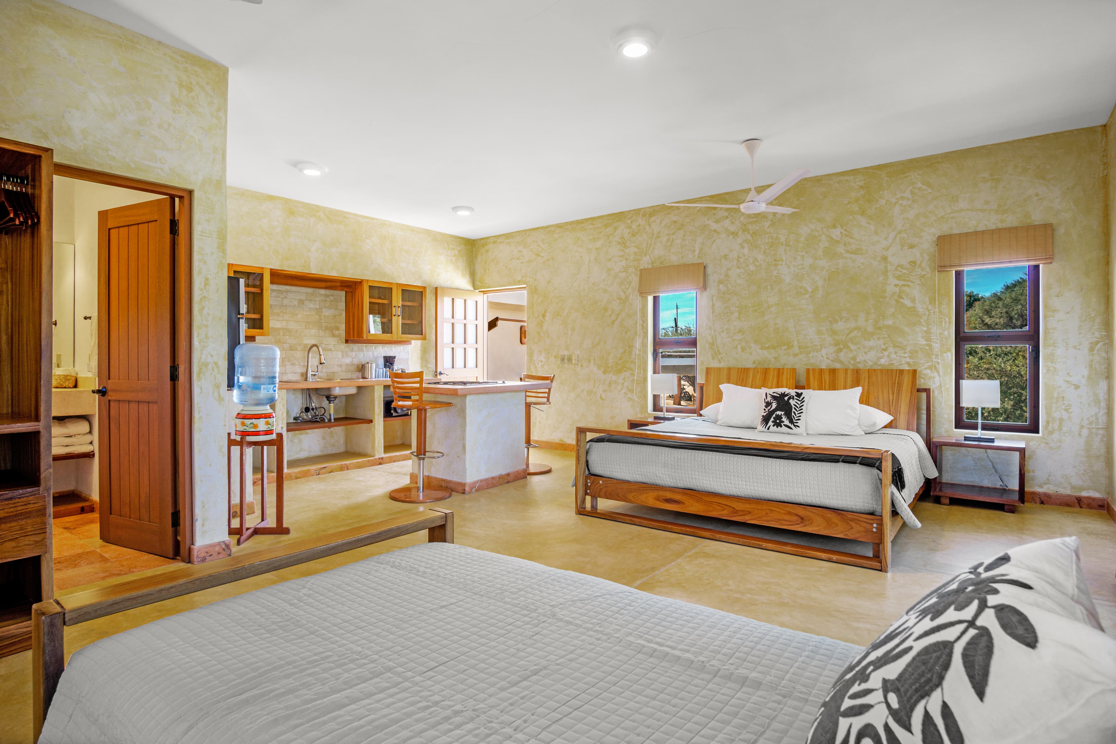 A spacious bedroom featuring two beds, a kitchen area, and large windows with natural light.