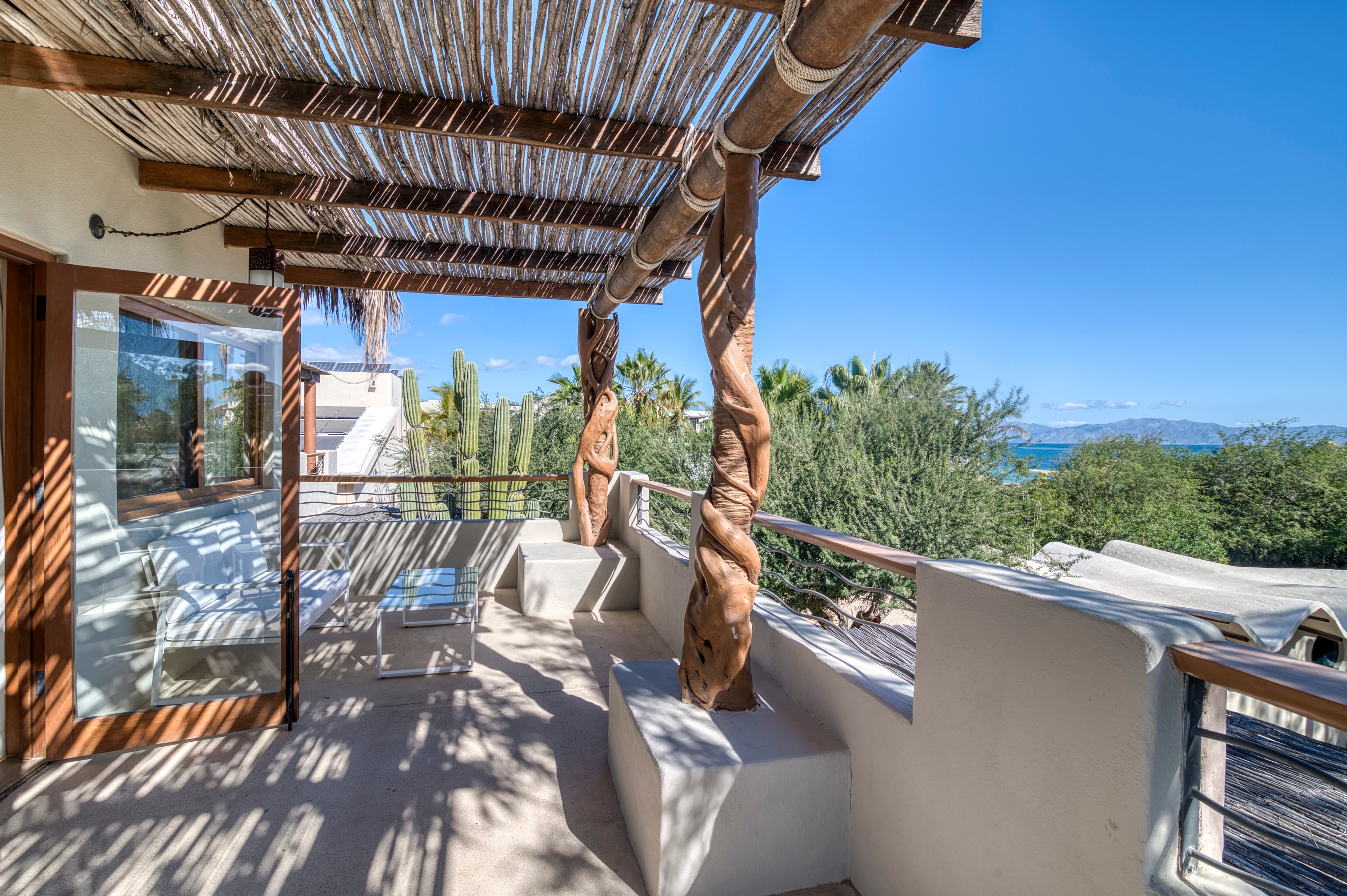 A sunlit balcony features wooden pillars, stylish seating, and a view of greenery and distant mountains.