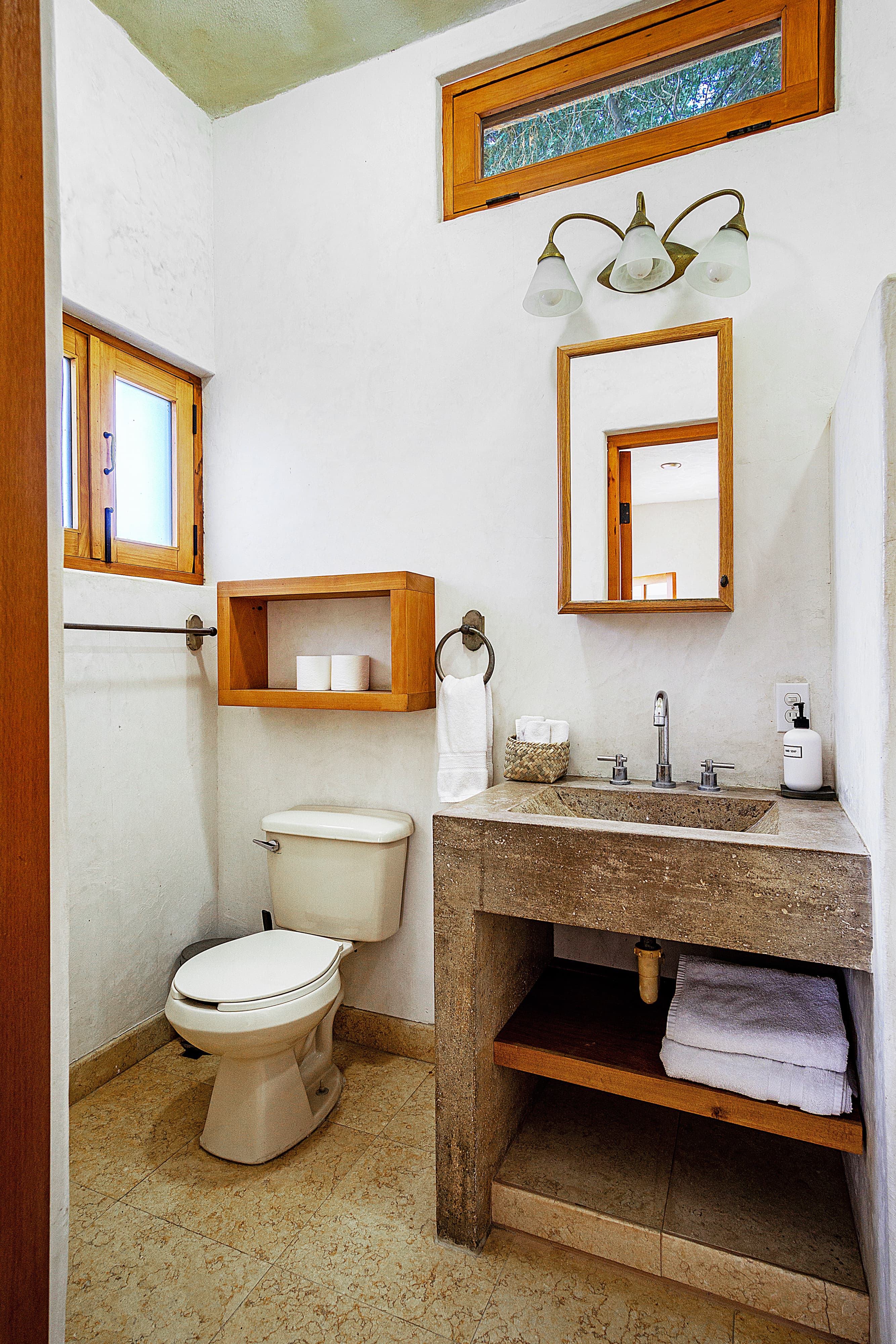 A modern bathroom featuring a toilet, stone sink, wooden shelving, and natural lighting from small windows.