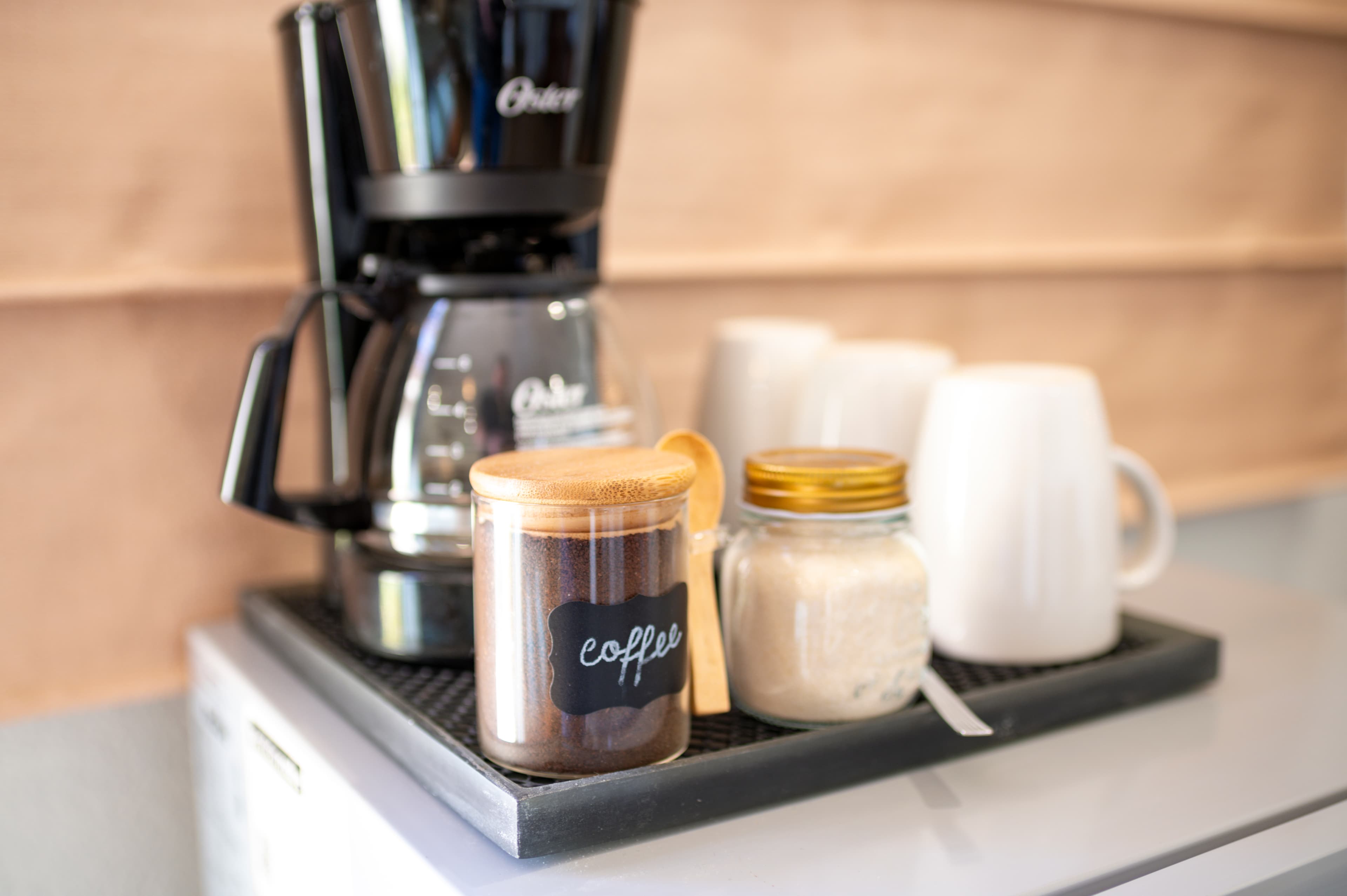 A coffee maker on a tray with jars of coffee and sugar beside white mugs.