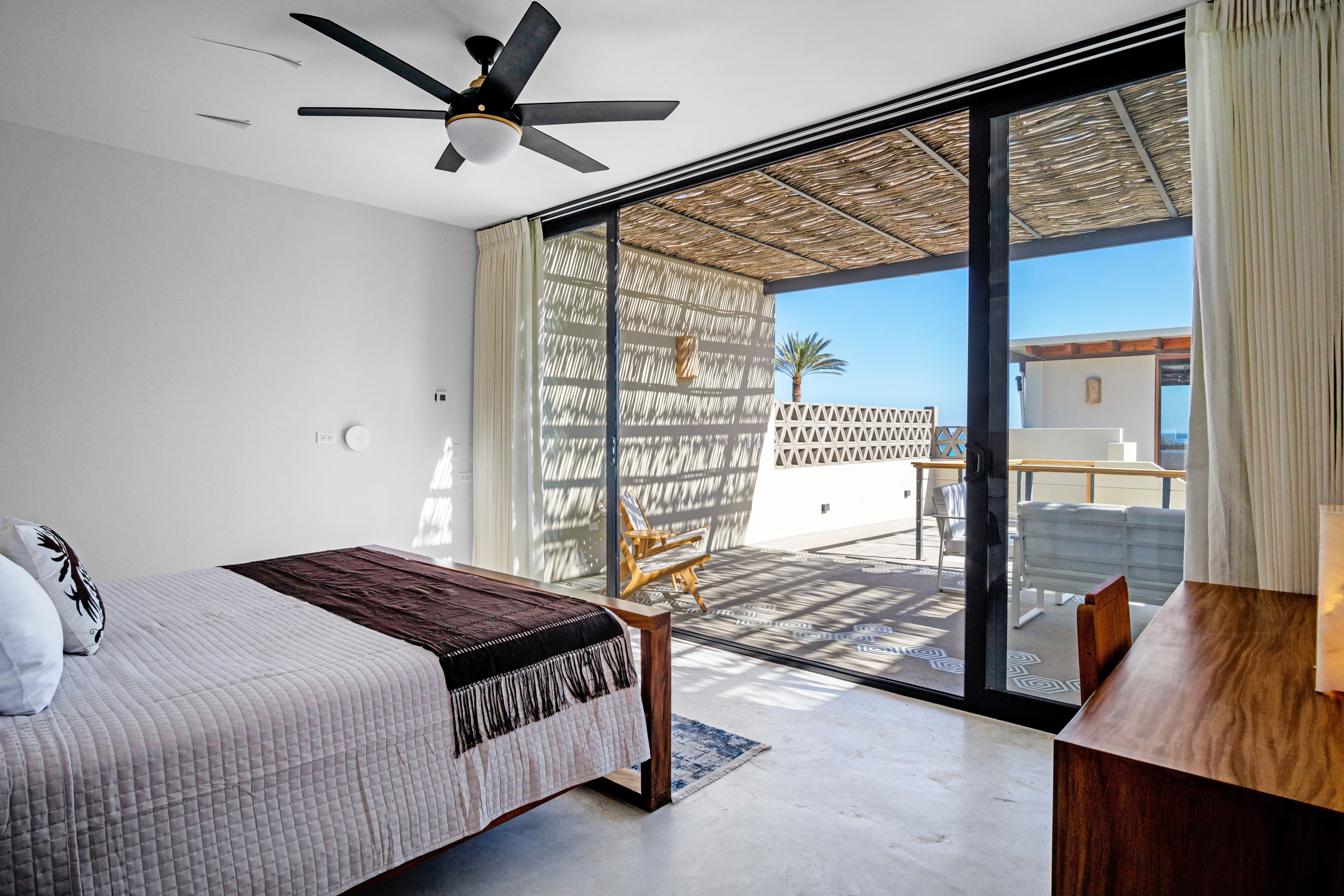 A modern bedroom with a bed and a view of a patio and ocean through large glass doors.