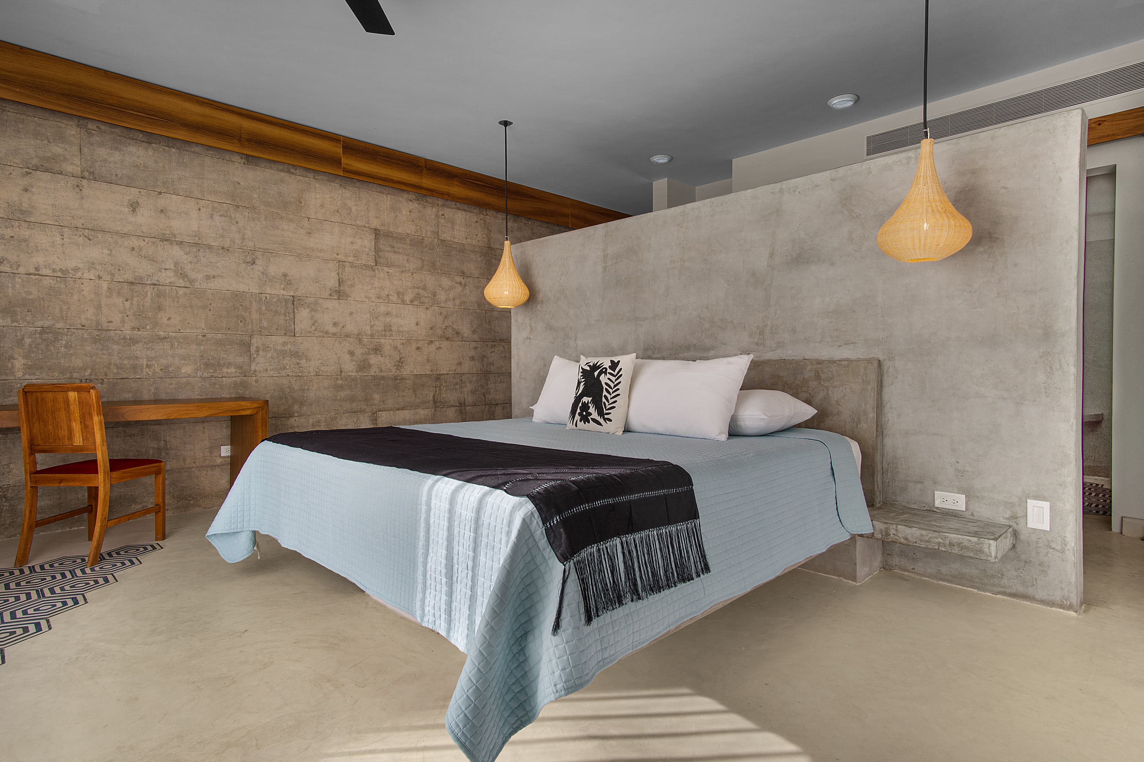 A minimalist bedroom featuring a bed with a light blue cover, white pillows, and concrete walls.
