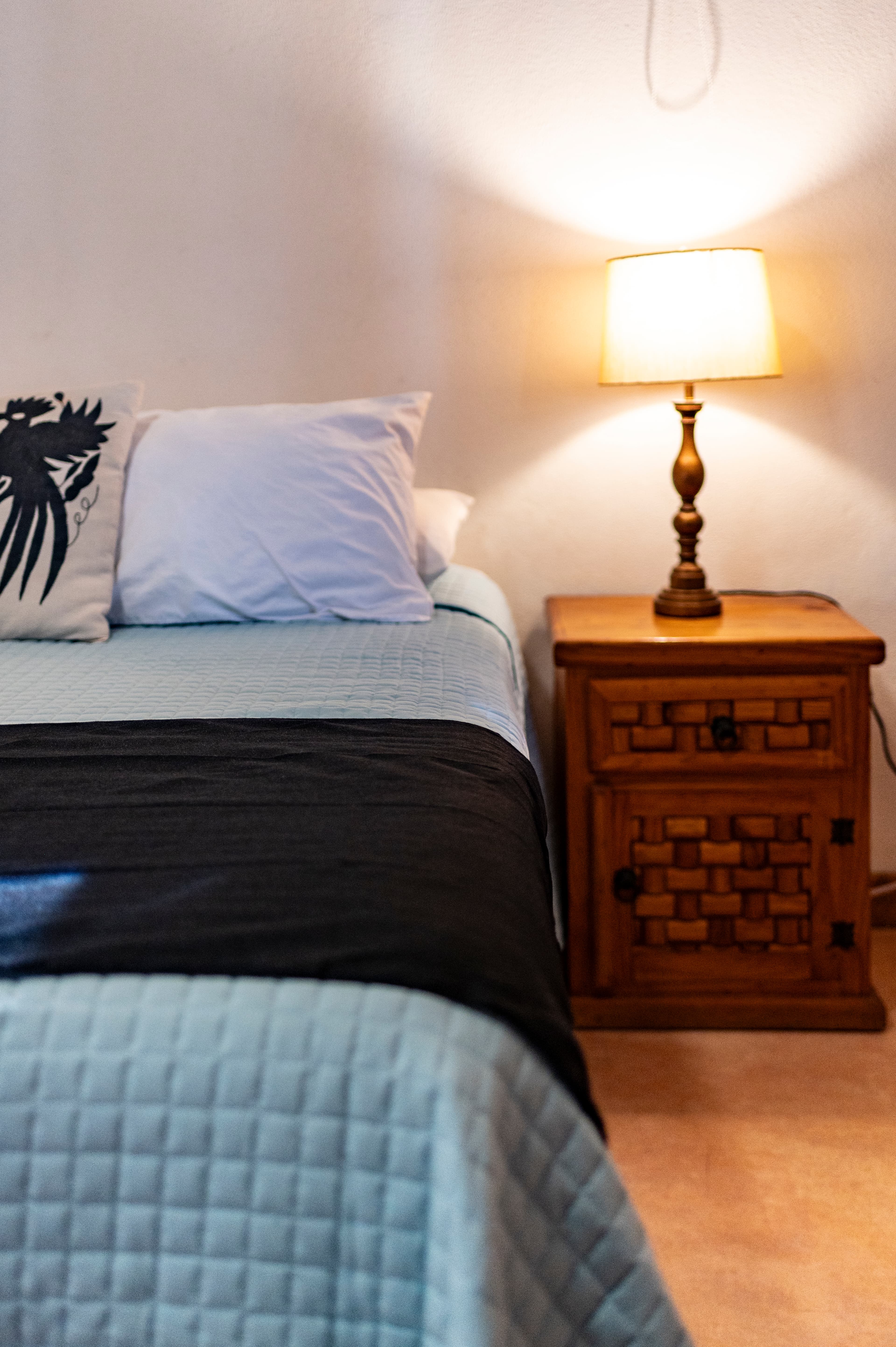 A cozy bedroom with a bed topped with pillows, a black throw, and a wooden nightstand beside a lamp.