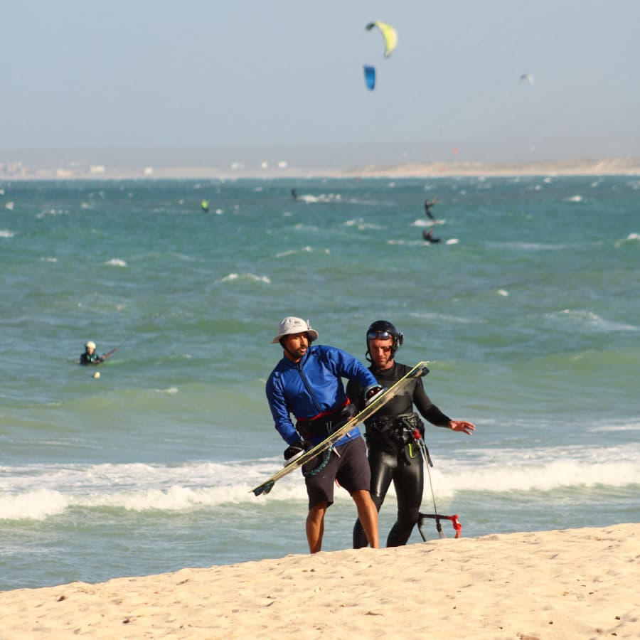Two kite surfers are walking along the beach, one carrying a surfboard while the other assists.