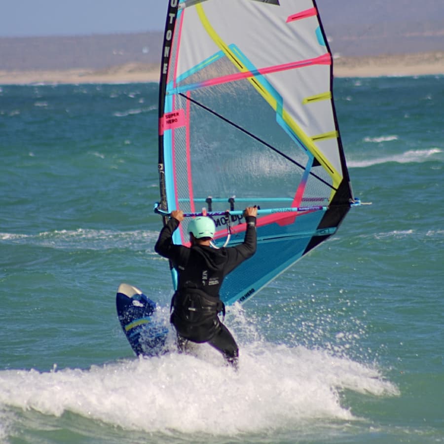 A person windsurfing on a vibrant blue sea, wearing a mint-green helmet and a wetsuit.