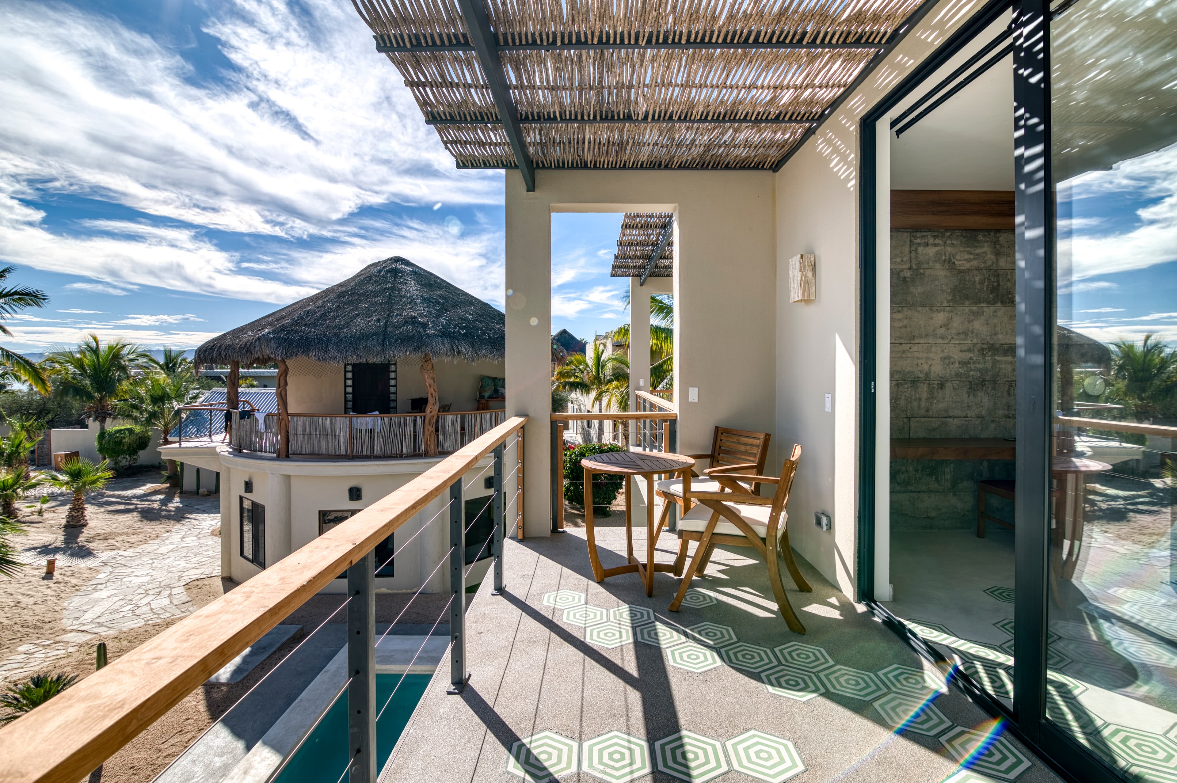 A wooden balcony with two chairs overlooks a tropical landscape featuring palm trees and a thatched-roof building under a blue sky.