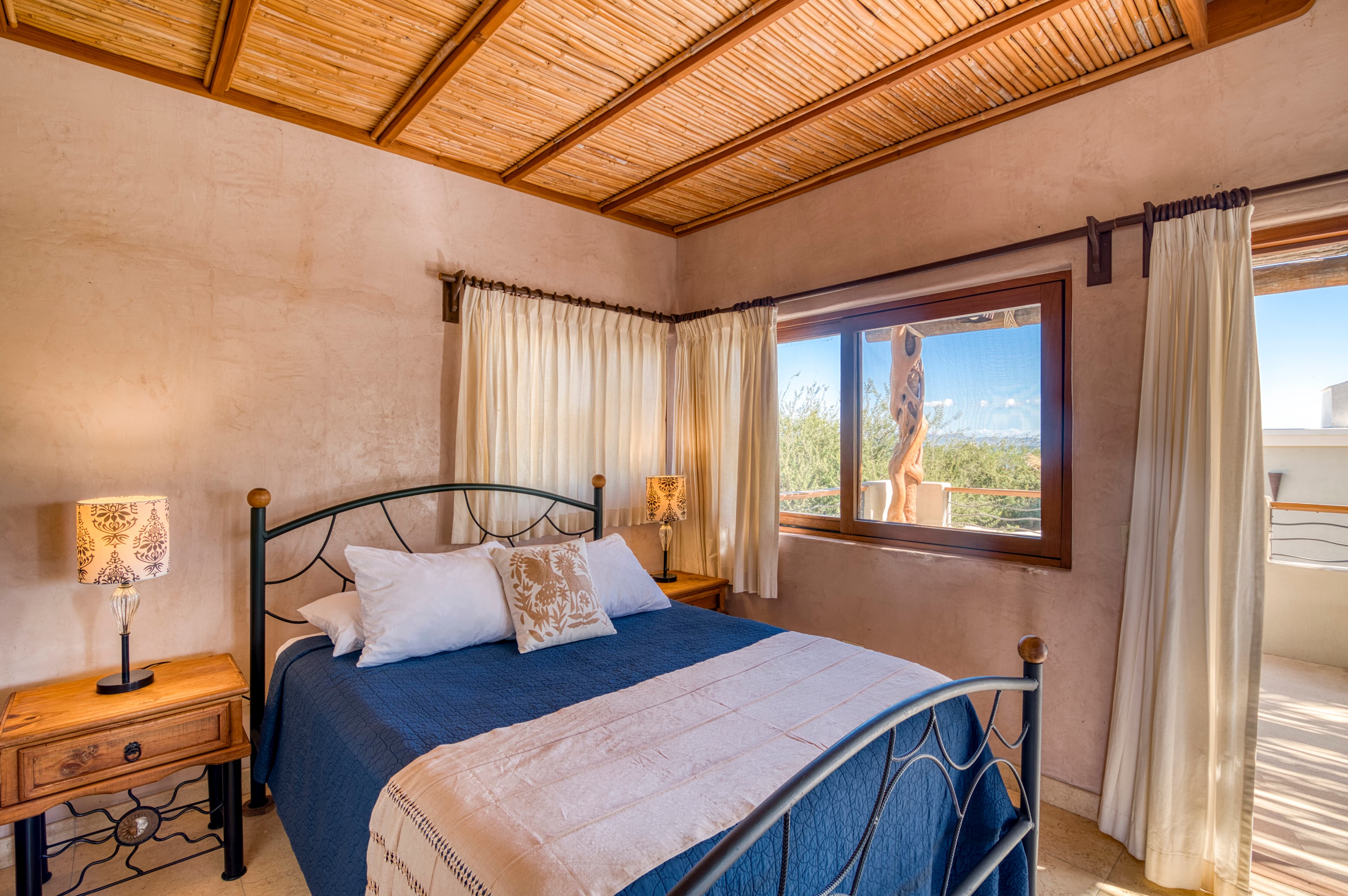 A cozy bedroom featuring a blue bedspread, wooden nightstands, and large windows with flowing curtains.